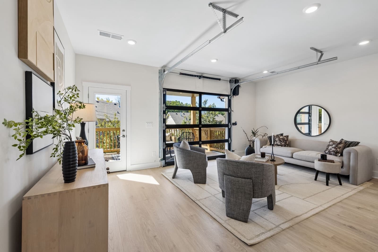 Living room with its black-framed glass garage-style door rolled up onto a covered deck at Sonya Park, with a glass side door to the deck at left and neighboring rooftops and trees beyond.