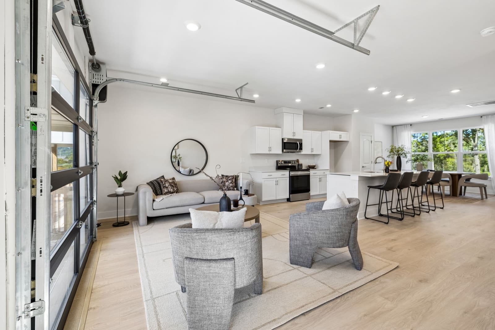 Open living, kitchen and dining level at Sonya Park with the glass garage-style door closed at left, white shaker cabinets, a kitchen island with four black stools and a dining table by a bank of windows.