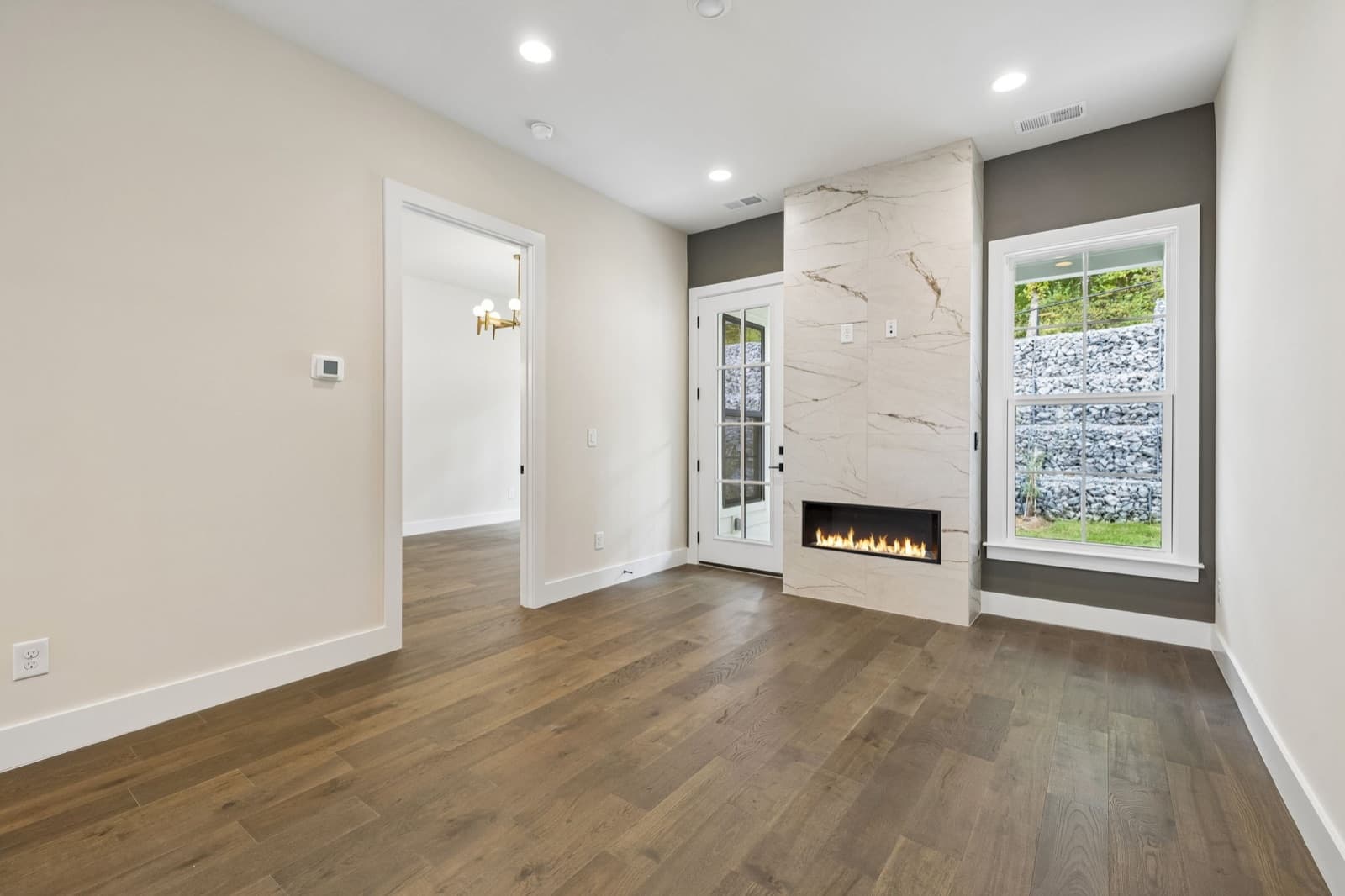 Living room at 117 Shadow Springs with a lit linear gas fireplace in veined tile, a glass door and a window onto the stacked-stone wall.