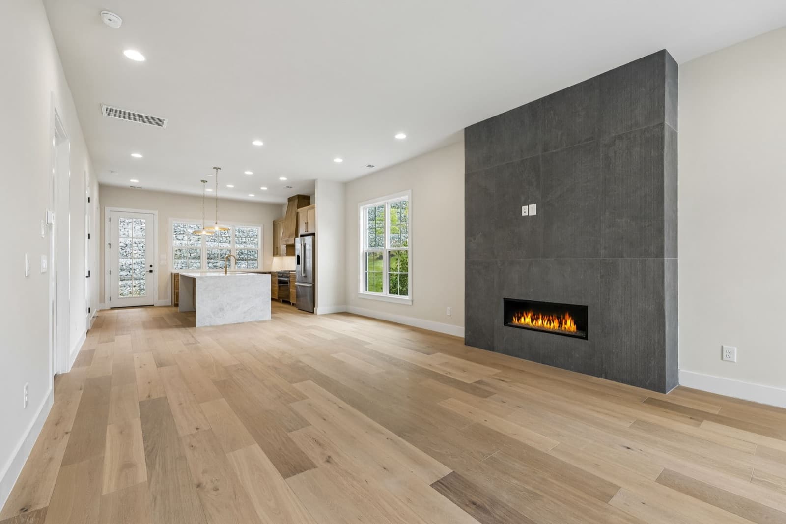 Living room at 125 Shadow Springs with a lit linear fireplace in a wall of dark tile, the kitchen island and windows onto the stone wall beyond.