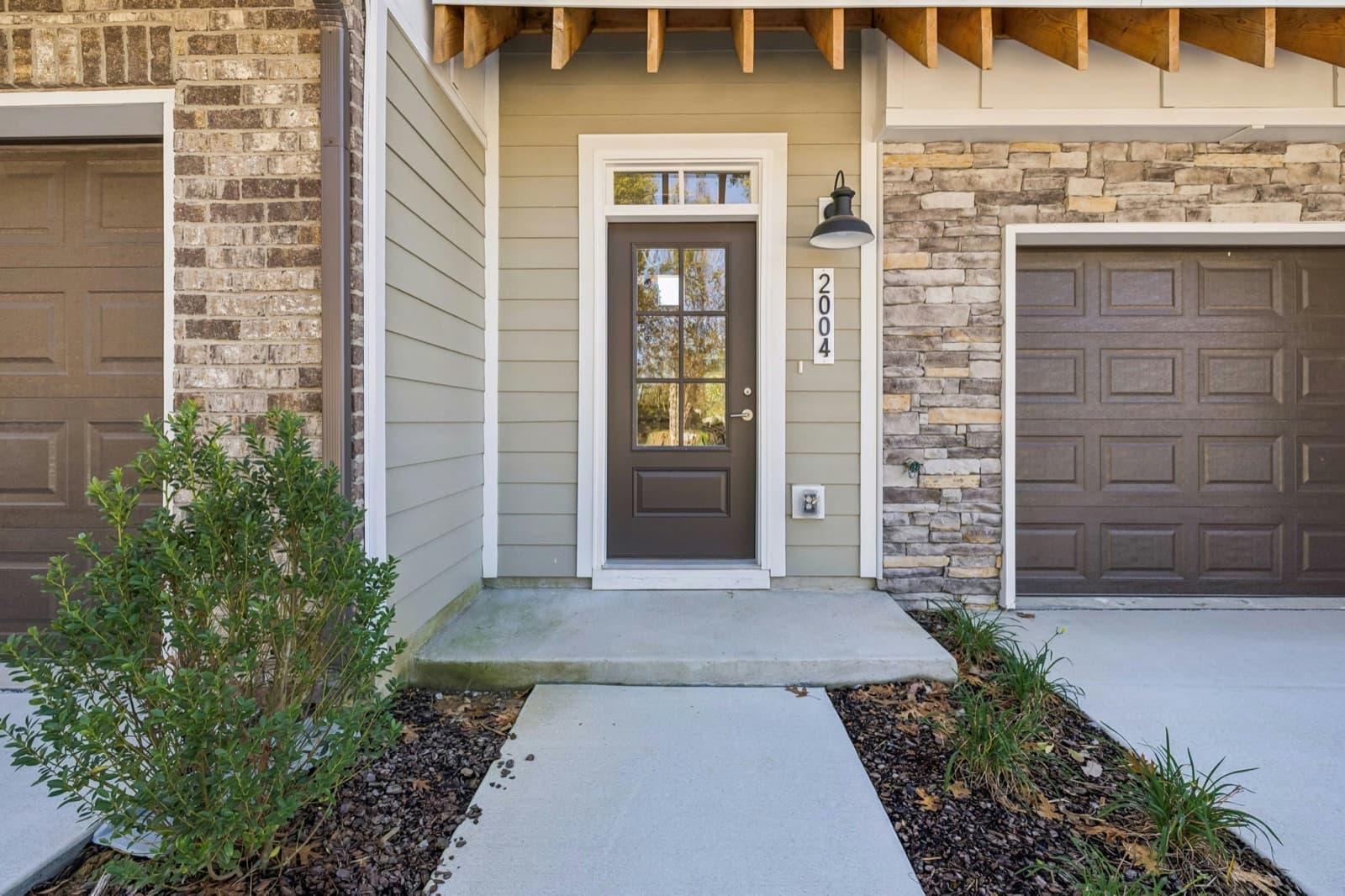 Front entry of 2004 Sonya Terrace: a dark glass-paneled front door with a transom, the house number 2004, a black gooseneck lantern and a timber-bracket overhang, with stacked-stone and brick garage fronts and dark brown garage doors on either side.