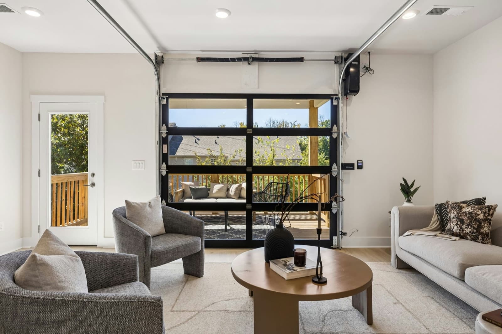 Furnished living room in the model at 2004 Sonya Terrace, looking straight at the closed black-framed glass garage-style door with deck furniture and a wood railing visible through the glass, a glass deck door at left, a round wood coffee table and gray chairs.