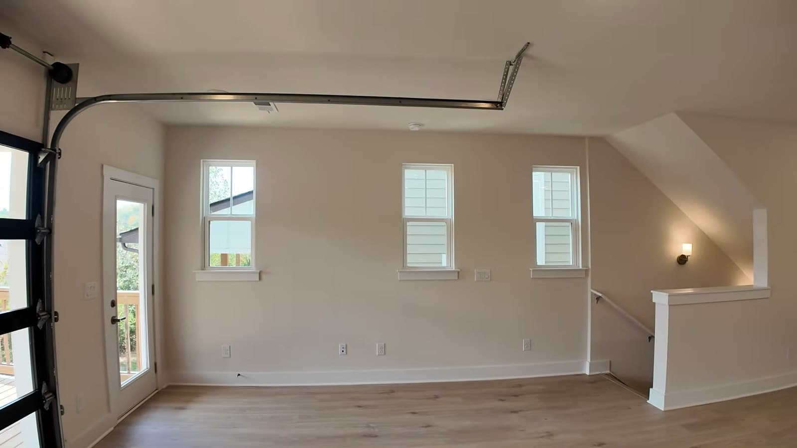 Empty living room at 2008 Sonya Terrace with three double-hung windows along the side wall, the overhead track of the glass garage-style door at the top left, a glass door to the deck and a stair rail at right, on wide-plank floors.