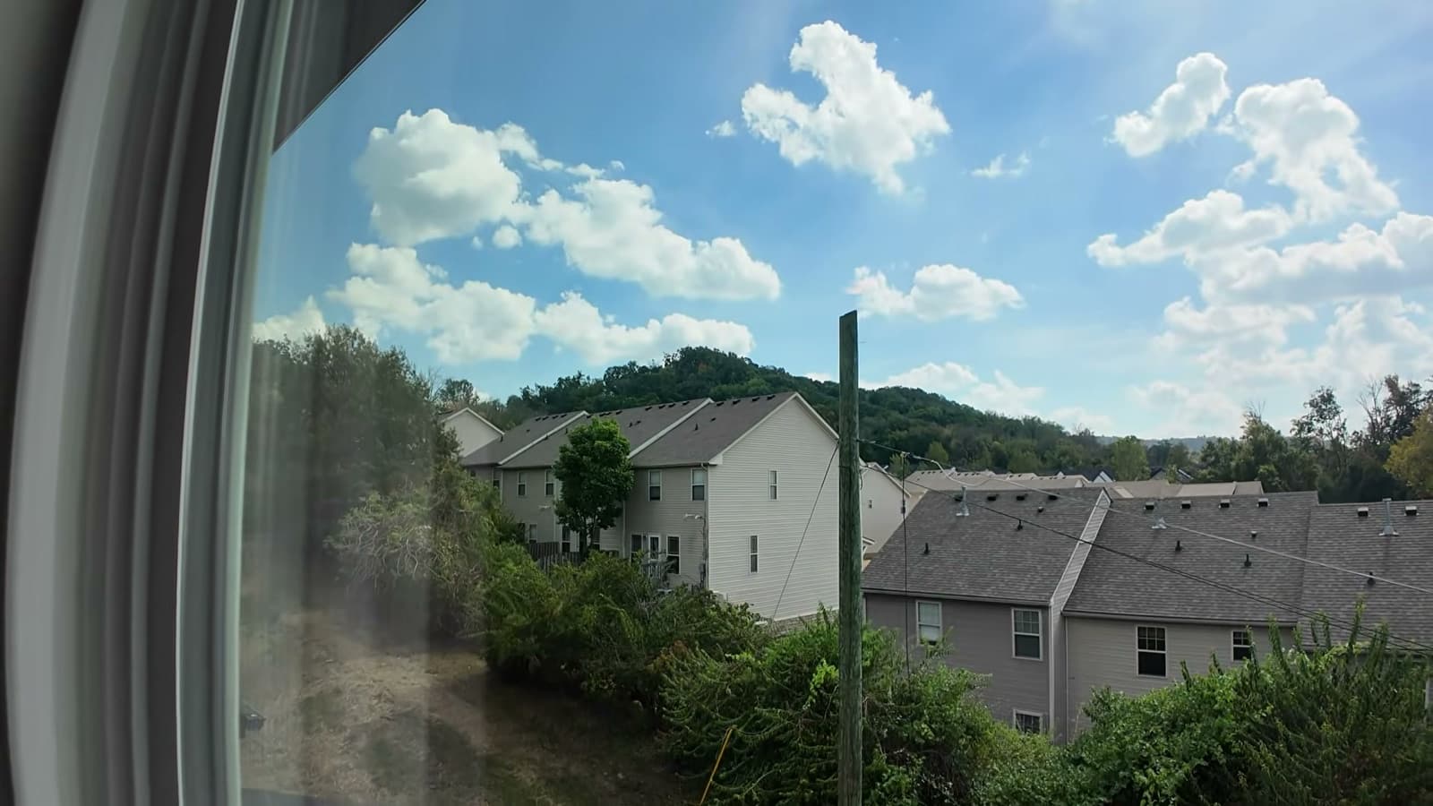 View from an upstairs bedroom window at 2008 Sonya Terrace: shingled rooftops of neighboring homes below, a wooded hill and a bright sky with cumulus clouds.